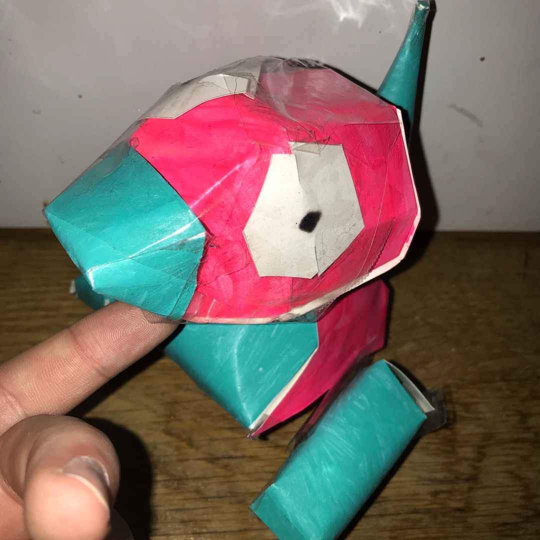 a paper figure of the pokemon porygon on a wooden floor with a white wall behind him. a finger is holding his head up