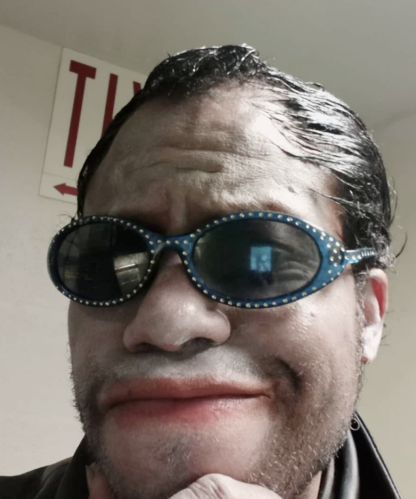 a man with slicked back black hair, his face is covered in white powder, and he's wearing messy red lipstick as well as blue bedazzled sunglasses. he's doing a smug face